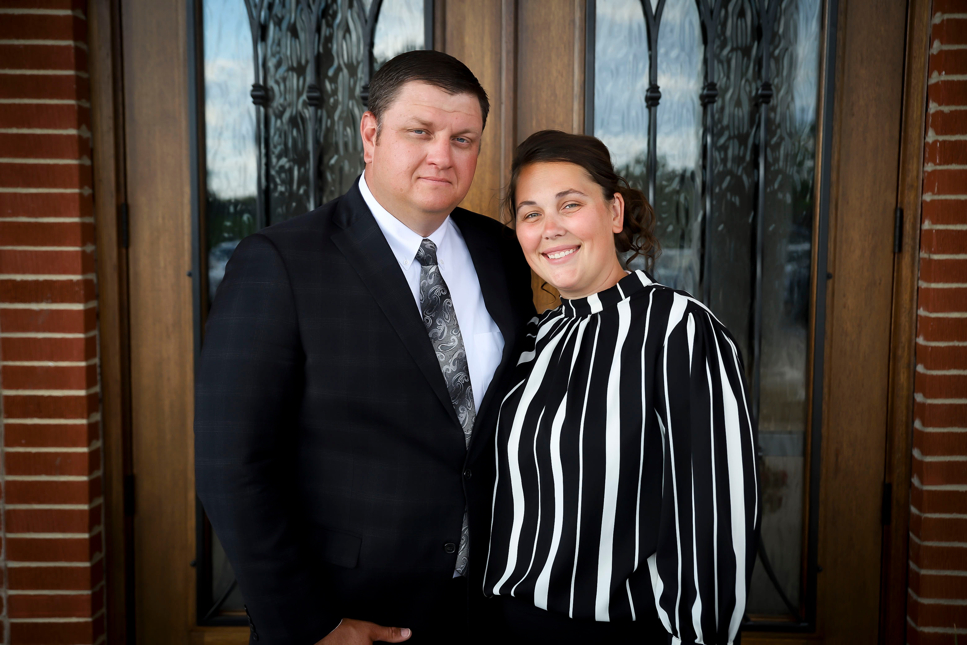 A man in a dark suit and a woman in a black and white striped blouse smiling together in front of the church's decorative glass doors.