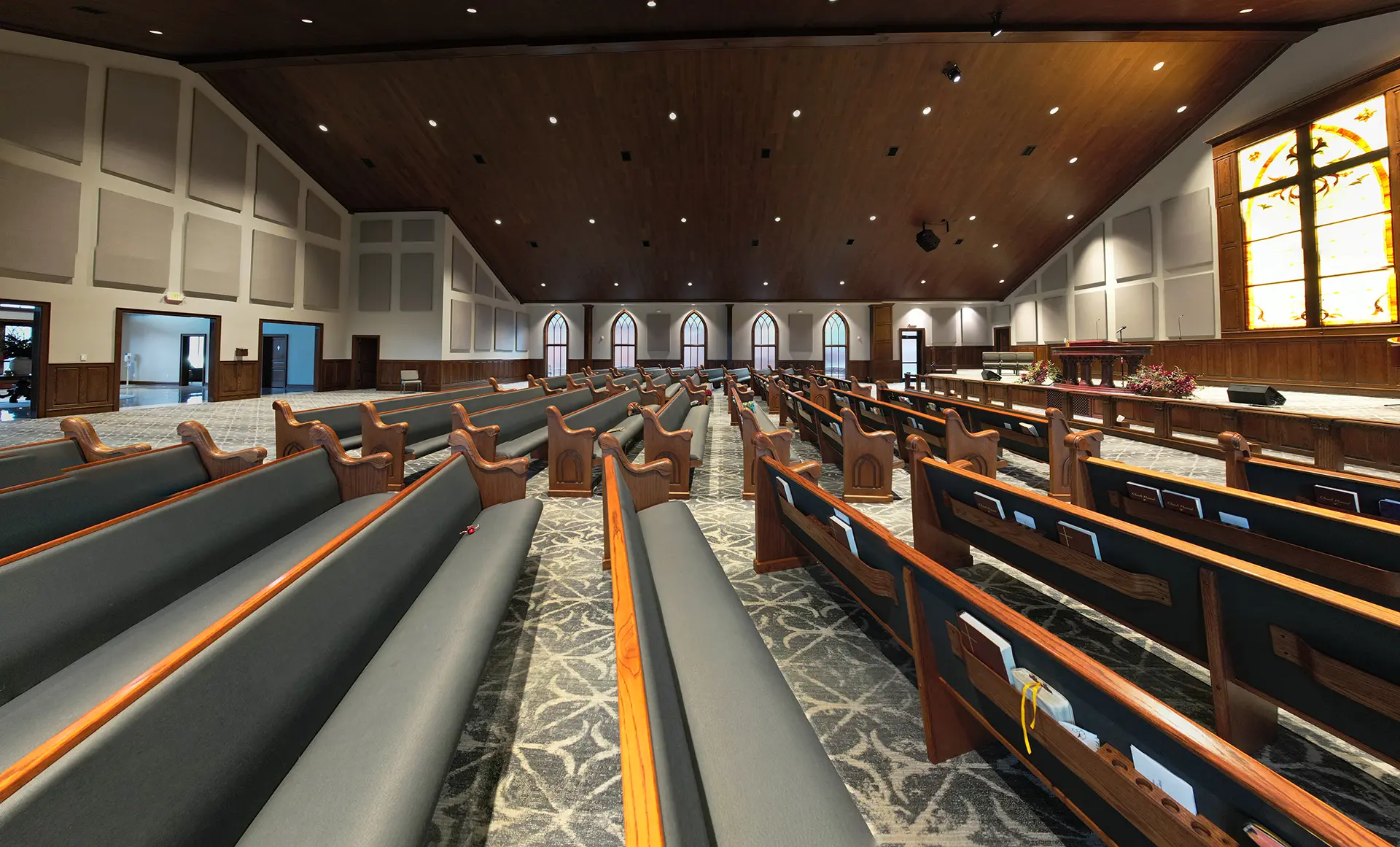 Interior of a modern church with rows of wooden pews, patterned carpet, and a large illuminated stained glass window at the front.